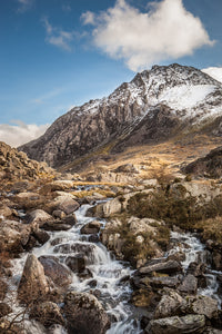 A photograph from below, looking upstream towards the imposing mountain of Tryfan. A lovely Winter's day with cloudy blue sky above the snow topped mountain. Ogwen Valley, Snowdonia, North Wales