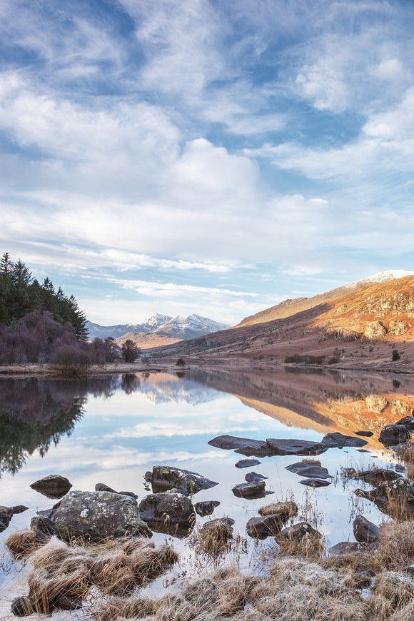 Llyn Mymbyr, Snowdonia - North Wales. An upright image with rocks in the foreground, looking across the still lake, reflecting it's surroundings, to Snowdon Horseshoe in the distance